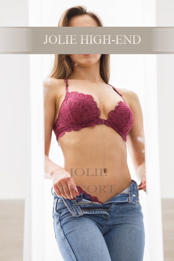 Highclass Escort in Lingerie mit Jeans, lächelnd, vor neutralem Hintergrund.