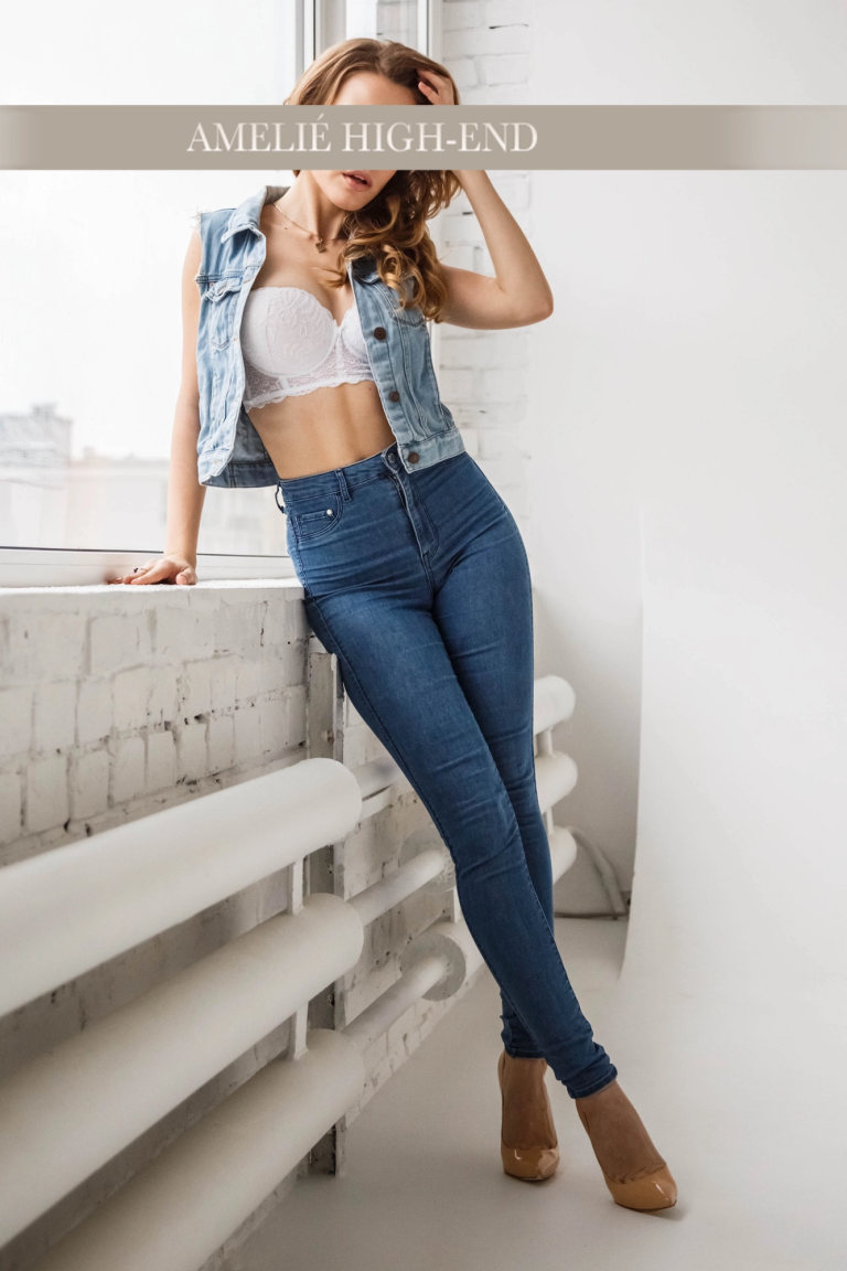 Escort Girl in Weste und enger Jeansposiert an einer Wand, lächelnd, mit hohen Schuhen.