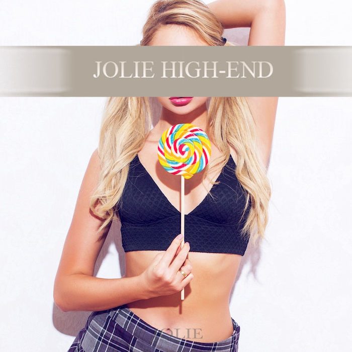 Escort Kati in einem schwarzen Top hält einen bunten Lutscher vor einem neutralen Hintergrund.