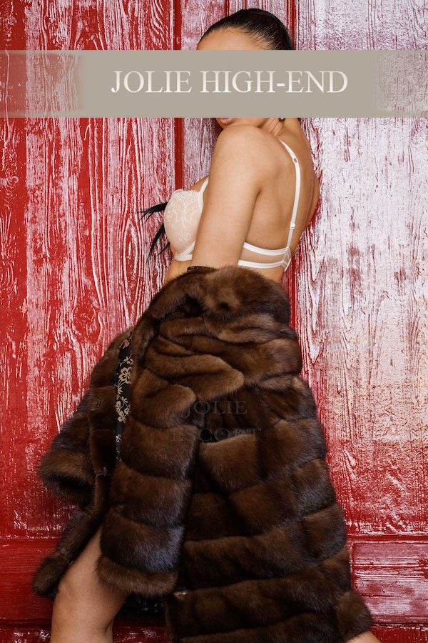 Hure Berlin in modischer, brauner Pelzjacke vor roter Wand.