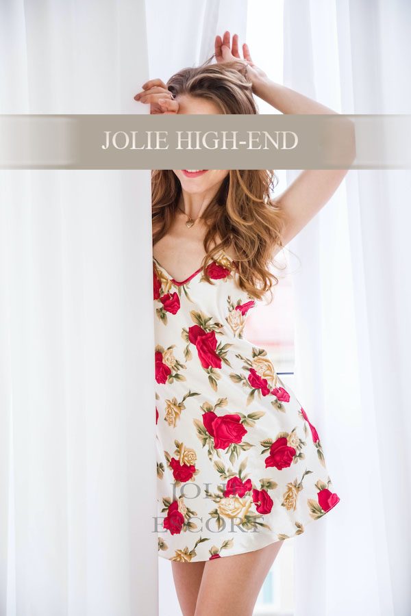 Escort Girl mit blonden Haaren trägt ein blütenverziertes Kleid hinter einem Vorhang.