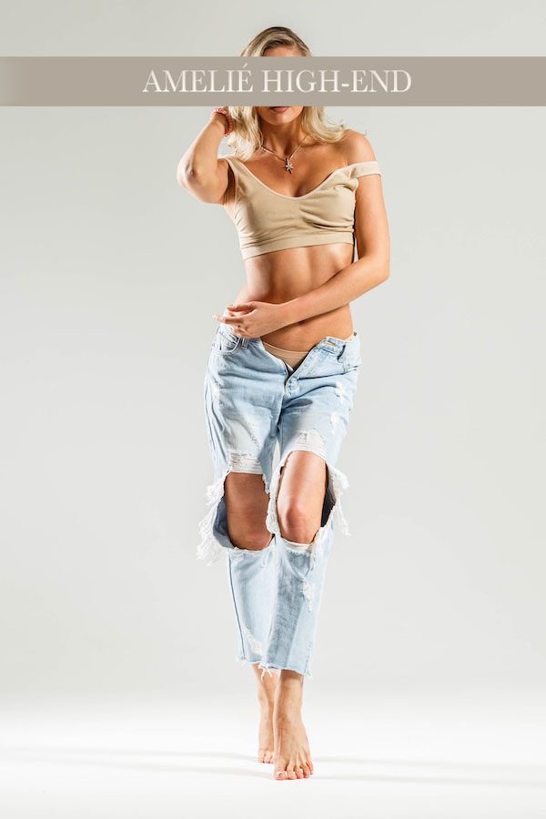 Junge Escort in beiger Top und zerrissenen Jeans, steht barfuß in halbseitiger Pose.