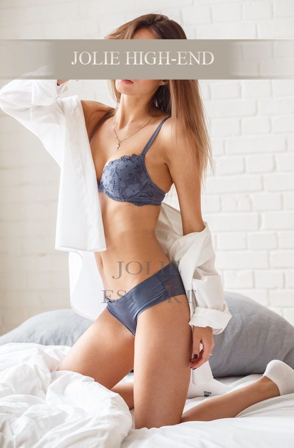 Frau in Unterwäsche sitzt auf einem Bett, trägt ein offenes Hemd.