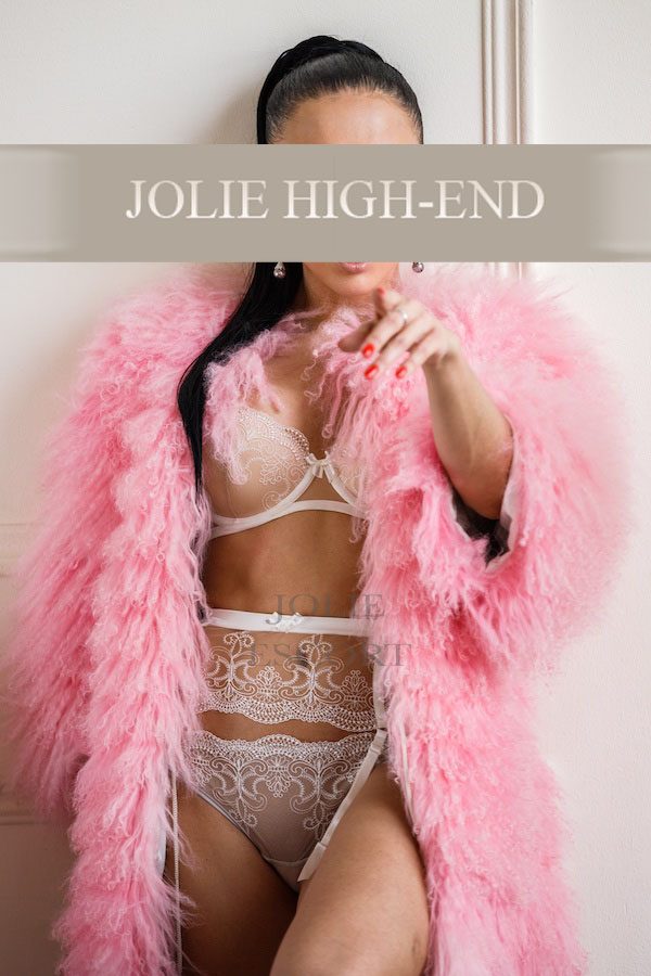 Highclass Escort in Berlin in einem pinken Pelzmantel und sexy Unterwäsche, mit den Händen auf die Kamera zeigend.