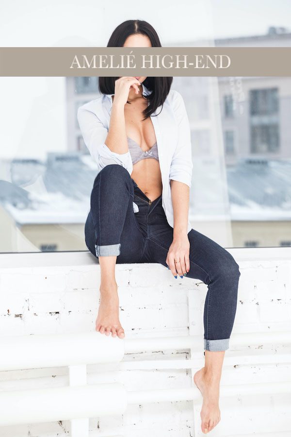 Escort sitzt in moderner Kleidung auf einem Fensterbrett, mit einem urbanen Hintergrund.