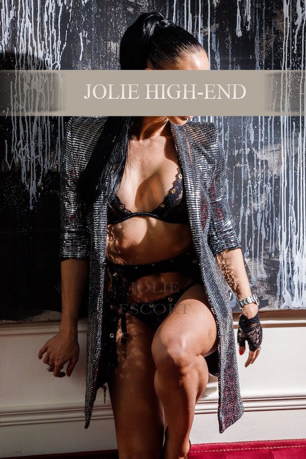 Devoter Escort Berlin in schwarzer Unterwäsche und glänzendem Mantel vor einem modernen Hintergrund.