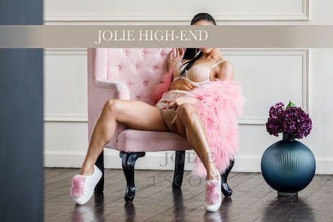 Highclass Escort Berlin in einem rosafarbenen Set sitzt auf einem pinken Sessel mit Blumen im Hintergrund.