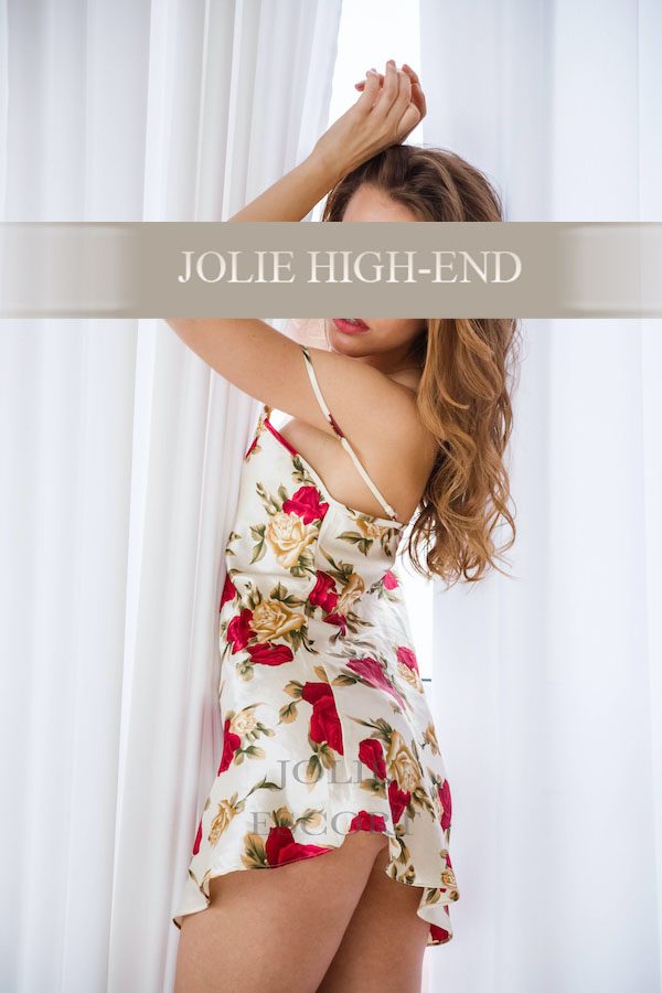 Escort Girl in einem floral gemusterten Kleid steht vor einem Fenster und posiert.