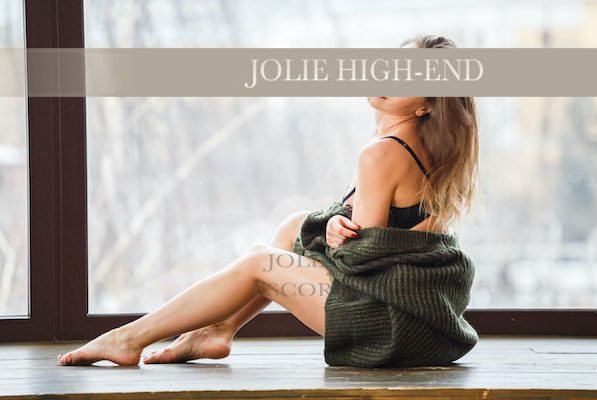 Eine Highclass Escort sitzt in einem Raum mit großen Fenstern, trägt einen Pullover und hat nasse Beine.