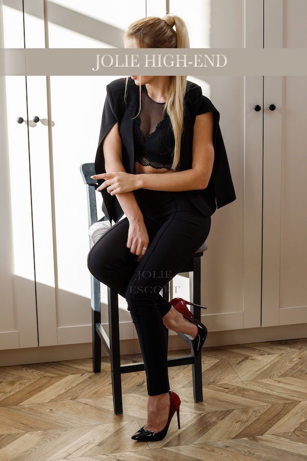 Nikki in schwarzer Kleidung sitzt auf einem Barhocker, Blick zur Seite gerichtet.