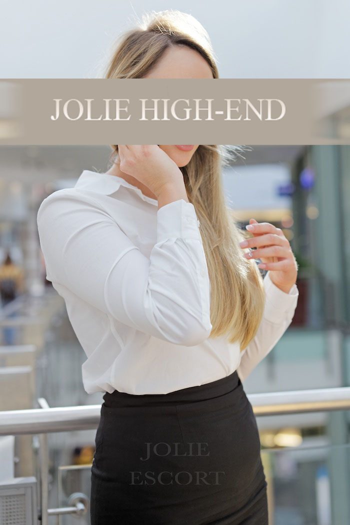 Blonder Escort Berlin mit blonden Haaren in weißem Hemd und schwarzem Rock, steht in einem modernen Raum.