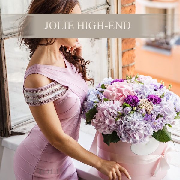 Escort Maggy in eleganter, rosa Kleid mit üppigem Blumenarrangement im Hintergrund.