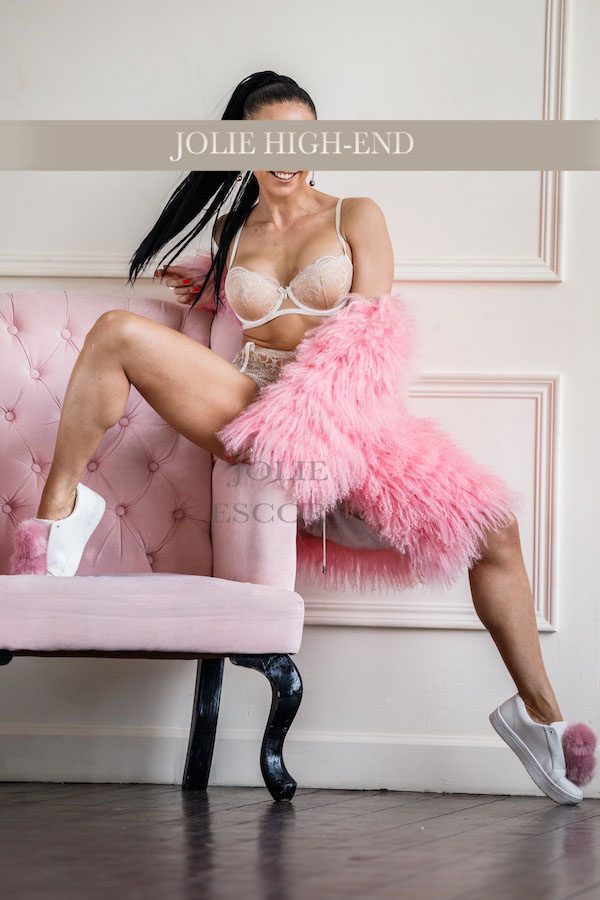 Begleitmodel in Berlin in Unterwäsche und rosa Pelzmantel sitzt an einem rosa Sofa.