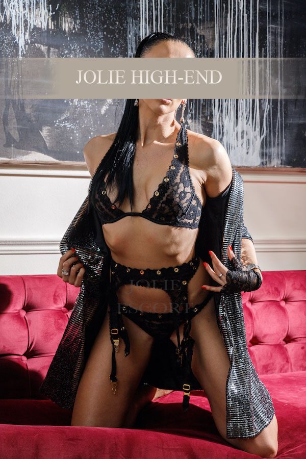 VIP Escort Berlin in schwarzem Spitzen-Bikini und elegantem Überwurf posiert auf rotem Sofa.