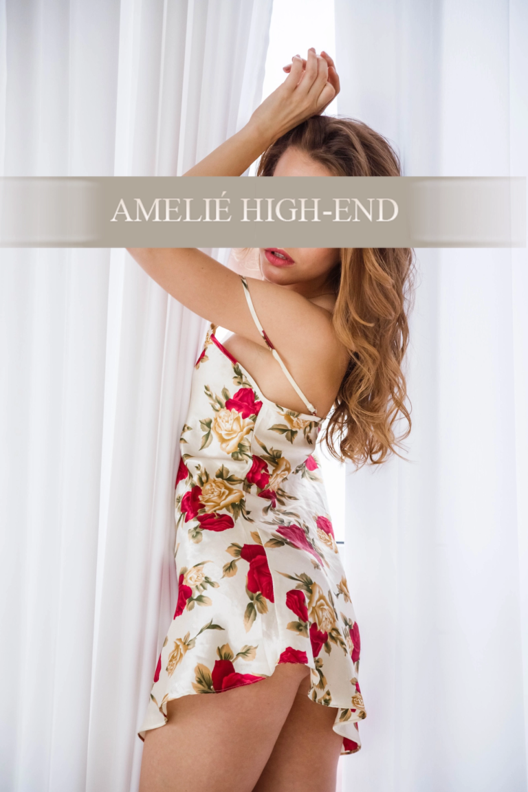 Escort Girl in einem floral gemusterten Kleid steht vor einem Fenster und posiert.
