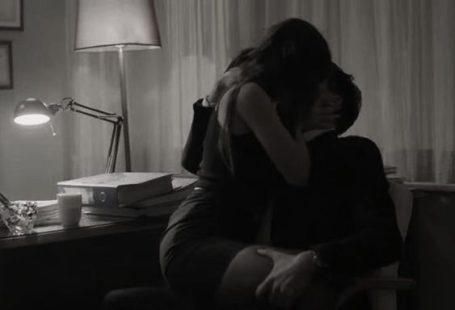 Paar in intimer Umarmung bei schwacher Beleuchtung, Tisch mit Büchern im Hintergrund.