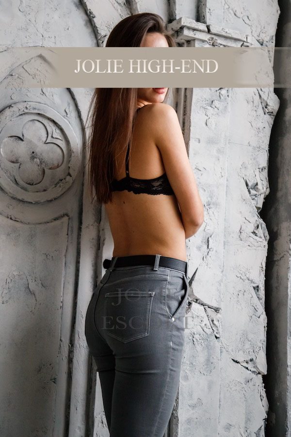 VIP Escort Frankfurt im schwarzen BH und Jeans, von der Seite mit angelehntem Rücken zur Wand.