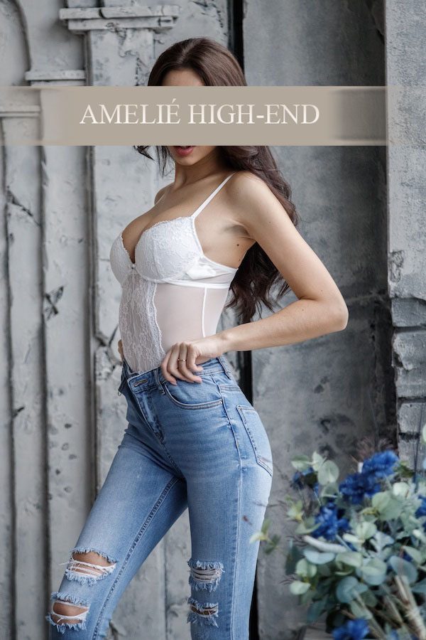 Escort in modischer Bluse und Jeans, posierend vor einer neutralen Hintergrundkulisse.