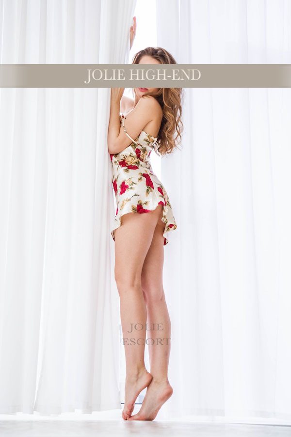 Escort Girl in floral Kleid, steht barfuß neben einem weißen Vorhang und lächelt.