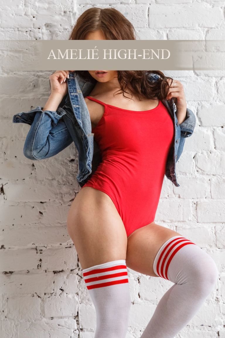 Escortmodel in rotem Einteiler mit weißen Strümpfen und Jeansjacke vor einer weißen Backsteinwand.