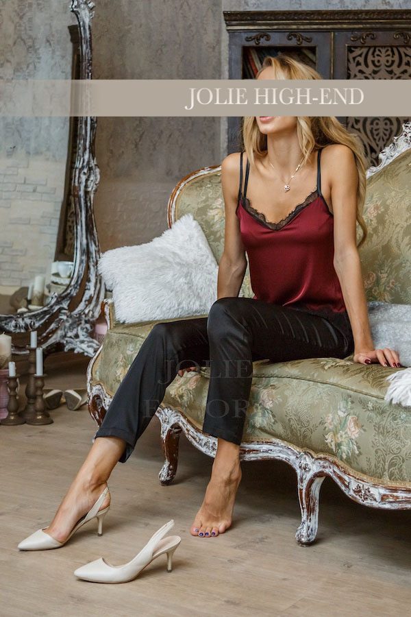 VIP Escort in rotem Top und schwarzen Hosen sitzt entspannt auf einer Couch.