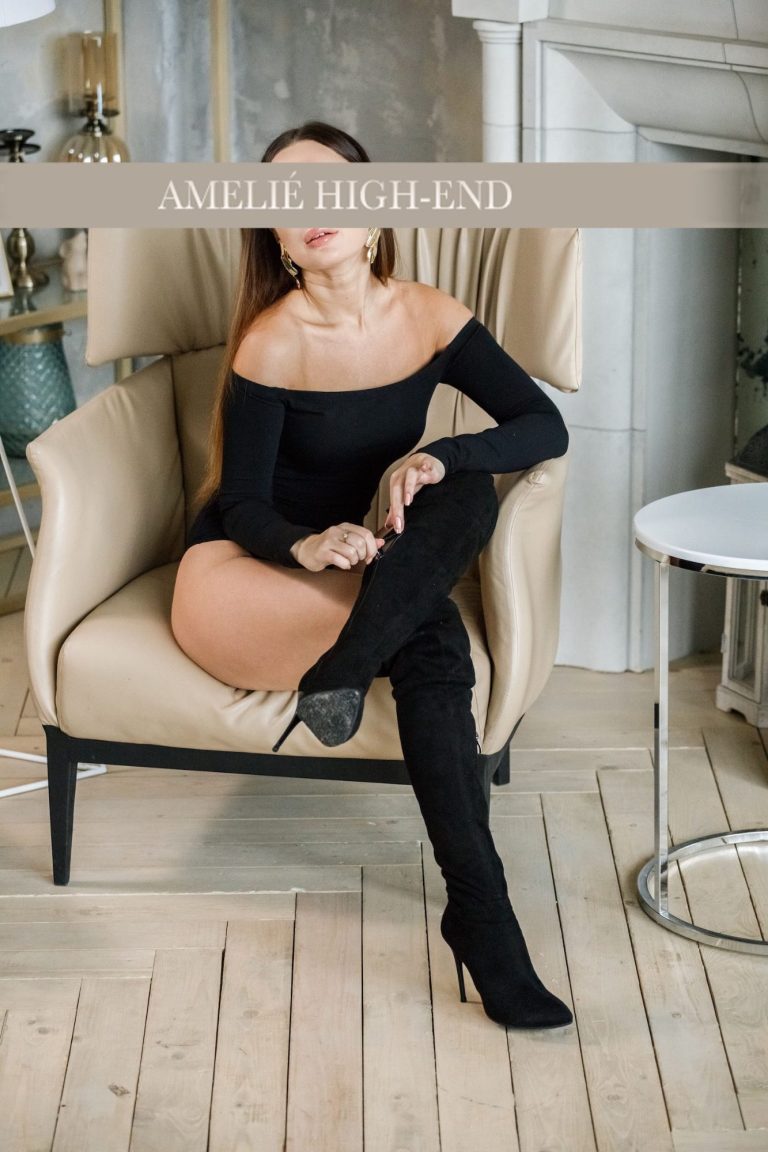 Escort Rebecca in schwarzem Kleid sitzt auf einem Stuhl mit gekreuzten Beinen.