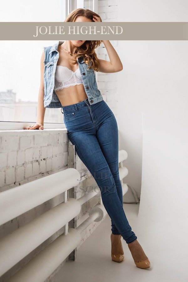 Escort Girl in Weste und enger Jeansposiert an einer Wand, lächelnd, mit hohen Schuhen.