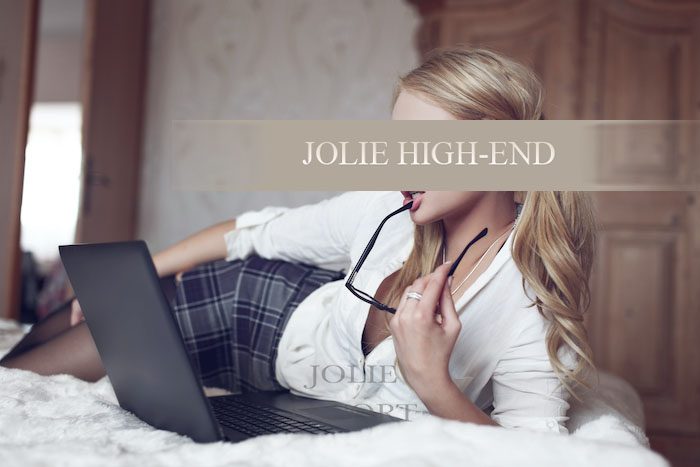 Escortservice mit Kati mit blonden Haaren sitzt auf einem Bett und hält eine Brille, während sie auf einen Laptop schaut.