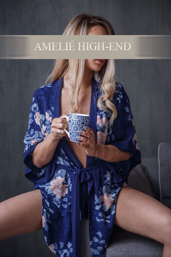high class escort in floral Kimono hält eine Tasse, sitzt entspannt mit leicht gewendem Blick.