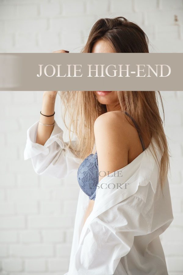 Schönes Escortgirl aus Frankfurt mit langen Haaren, trägt ein Hemd und einen BH, vor einer weißen Wand.